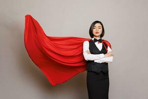 Confident asian waitress standing with folded arms and posing in fluttering superman red cape. Young attractive woman receptionist dressed in hero cloak and catering service uniform photo