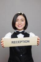 Cheerful asian restaurant waitress showcasing stainless steel reception plate portrait. Smiling hotel administrator showing receptionist name signage and looking at camera photo