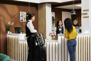 Friendly hotel staff helps client settle in, offering professional support and preparing to take her baggage to room. Asian concierge employee and customer having pleasant talk in foyer photo
