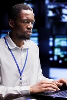 African american IT developer in data center using laptop to set up configuration management tools that enable automatic failover and load balancing, preventing server cabinets system crash photo