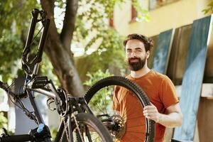 Portrait shot of energetic male cyclist holding dismantled bicycle tire for repair and maintenance in home yard. Active sporty caucasian man handling bike wheel for reattachment. photo