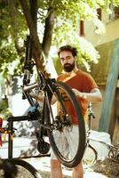 imagen exhibiendo comprometido sano atlético hombre meticulosamente inspeccionando moderno bicicleta para regular verano mantenimiento. activo joven caucásico masculino cuidadosamente avaro bicicleta componentes para reparar. foto