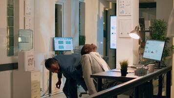 African american accountant sneaking up on sleepy coworker napping at desk during nightshift. Funny office clerk playing joke on sleeping colleague, adjusting his chair height level from behind photo