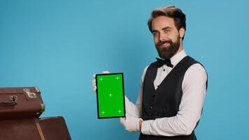 Stylish bellboy presents tablet in studio and showing blank greenscreen display while he stands next to trolley bags. Classy porter showcasing concierge assistance with luxury service. photo