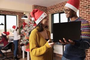 Smiling diverse colleagues laughing while doing teamwork on laptop in workplace at christmas eve. Man and woman employees in santa hats holding portable computer in decorated office photo