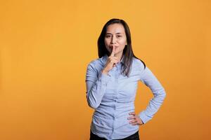 Portrait of filipino woman making silence gesture by putting forefinger over lips posing over yellow background. Silent woman shushing people indicating secrecy and confidentiality. photo