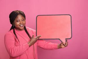 Portrait of positive cheerful woman holding red cardboard speech bubble standing in studio on pink background. Joyful young adult holding blank template announcement placard. photo