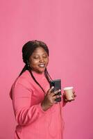 Smiling woman with pink sweather holding cup of coffee while reading the news on phone, posting picture social media. Young adult watching funny video on smartphone whle posing in studio photo