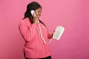 Portrait of cheerful young adult chatting on landline phone line discusssing with friend while standing in studio with pink background. Woman having remote conversation on stationary vintage phone photo