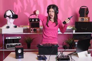 Unhappy performer holding microphone while doing thumbs down gesture, expressing disagreement in studio over pink background. Musician performing song with electronics equipment and audio instrument photo