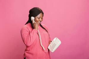 retrato de sonriente africano americano modelo chateando en teléfono fijo teléfono línea mientras en pie en estudio con rosado antecedentes. mujer teniendo remoto conversacion en estacionario Clásico teléfono foto