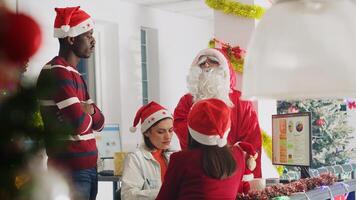 Revealing shot of manager dressed as Santa during Christmas season delegates project tasks to workers. Management executive in xmas decorated workspace discussing with employees photo