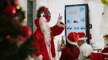 Manager dressed as Santa during Christmas season holding team meeting, showing next year company strategy on digital screen. Team leader in festive decorated office talking with employees photo
