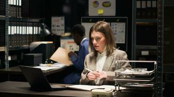 Detailed view of female investigator entering office and sits to use laptop for forensic research. African american detective and caucasian officer investigate a case, going through files and evidence photo