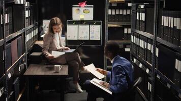 In room crammed with clues and records, caucasian female investigator sits on table analyzing evidence with police officer. African american inspector hands over case data to policewoman with laptop. photo