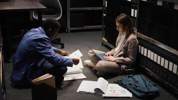 Multiethnic detectives sit on floor of archive room conducting investigation while surrounded by papers, piles of evidence, and folders. Using laptop, female investigator gathers case material. photo