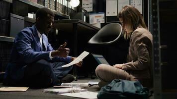 Male inspector and female police officer seated on office floor with paperwork and laptop, conducting crime investigation. Case clues and evidence are examined and discussed by multiracial detectives. photo