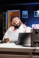 Detailed shot of Arab man using a laptop and mobile device for communication and research, showcasing proficiency in technology. Muslim guy multitasking, talking on his cellphone and comparing notes. photo