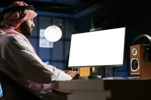 The image depicts A Muslim man sits at his home workstation, his computer monitor displaying an isolated chromakey template. Young Arab man working on a desktop computer with a copyspace white screen. photo