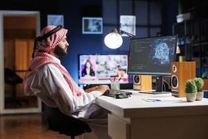 A professional man dressed in Arabic clothing, is working on futuristic interface while seated at his home office desk. Muslim guy manages data, monitors security, and programs AI on desktop computer. photo