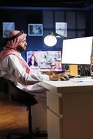 Image shows an Islamic man sitting at his home office desk with the desktop pc monitor displaying a blank copyspace template. Dedicated Arab guy using the computer having an isolated white screen. photo