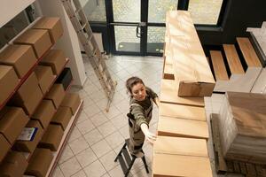 Top view of storehouse worker standing on ladder, checking cardboard boxes full with merchandise. Storage room supervisor preparing customers order before shipping products in warehouse photo