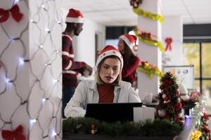 Fatigued employee struggling with overdue task deadlines in festive themed office while joyful coworkers have fun in background. Businesswoman working in Christmas decorated workspace photo