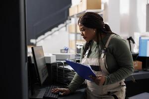 Stockroom supervisor looking at inventory report on computer, working at merchandise quality control in warehouse. African american employee preparing customers orders for shipping in storehouse photo