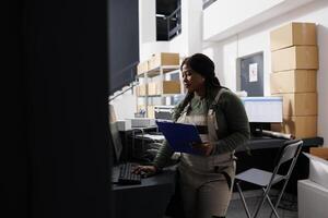 Storage room employee working at goods inventory, analyzing merchandise checklist on computer in storehouse. African american worker preparing customers orders during warehouse inventory photo