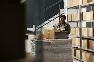 Stockroom worker preparing customers orders, using adhesive tape to pack products in cardboard boxes in warehouse. Storage room supervisor wearing industrial overall during storehouse inventory photo