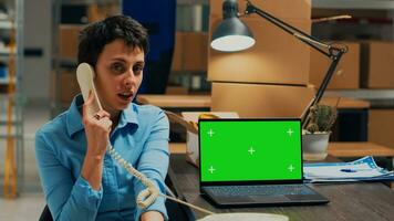 Young adult answering office telephone at desk with greenscreen, using chromakey isolated display. Warehouse employee planning shipment on landline phone call with blank mockup. photo