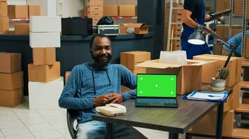 African american man using laptop with greenscreen template, planning merchandise shipment in warehouse. Male employee working with isolated display and chromakey copyspace screen. photo