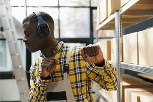 African american handyman with closed eyes dancing in warehouse. Storehouse loader wearing headphones listening to music playlist and making arms movements near shelf full of cardboard boxes photo