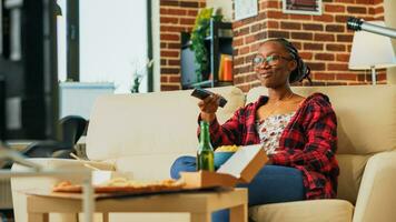 African american girl opening television to watch movie, eating chips aand drinking alcohol on couch. Smiling woman feeling happy with fast food meal in front of tv. Handheld shot. photo