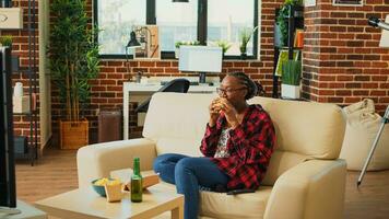 Young woman enjoying cheeseburger and beer at home, ordering fast food takeaway meal and watching favorite tv show. Cheerful person eating burger and fries, drinking alcohol at television. photo