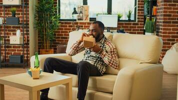 Modern man eating hamburger with fries on couch, having dinner with fast food and beer bottle at home. Young guy feeling relaxed watching comedy or action movie, having fun with tv. photo