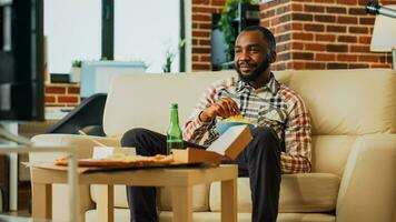 African american man eating chips in bowl at home, laughing at movie on television. Happy adult watcching tv series with snacks, bottle of beer and multiple food from delivery. Handheld shot. photo