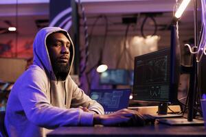 Hacker in hood hacking computer system and looking at camera. Young african american man breaking law and breaching data while working late in abandoned warehouse portrait photo