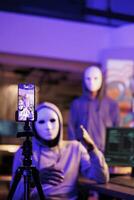 Smartphone screen with hackers recording threat video, asking victim for sensitive information and money. Criminals wearing anonymous masks threatening online and streaming fraud at night time photo