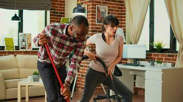 Diverse couple showing dance moves and having fun cleaning apartment, using mop to sweep dust and vacuum at home. Life partners dancing and singing, using washing solution, spring cleaning. photo