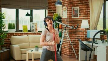 Positive woman listening to music on headphones and washing apartment floors, sweeping dirt. Housewife feeling happy cleaning household with mop and appliances, dance moves. photo