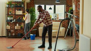 Interracial couple vacuuming floors and cleaning furniture on shelves, sweeping dust or dirt with tools. Life partners using vacuum cleaner with hose, gloves and rags to clean household. House chores. photo