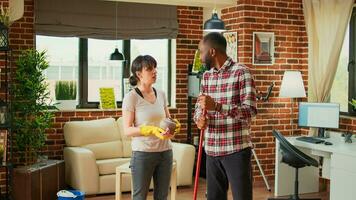 Young couple cleaning living room with vacuum and furniture polish, woman using all purpose cleaner to sweep dust from shelves. African american man vacuuming wooden floors at home. photo