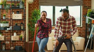 Happy man and woman showing dance moves and doing spring cleaning, having fun with music cleaning floors with mop and vacuum cleaner. Young smiling couple doing chores. Handheld shot. photo
