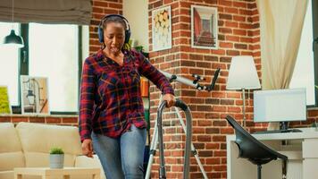 African american woman listening to music on headset and vacuuming floors in living room, using vacuum cleaner and having fun spring cleaning. Modern adult dancing and singing. Handheld shot. photo