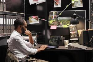 Private detective wathing cctv camera photos on laptop while sitting at workplace desk. African american investigator analyzing surveillance photographs on computer screen
