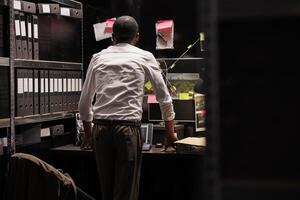 Detective analyzing crime weapon and photos attached to wall, searching insight. African american police investigator looking at evidence and photographs connection map at board