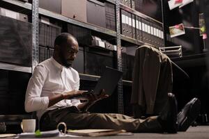 African american policeman sitting on floor and using laptop. Criminologist studying crime case file, checking information on portable computer in dark office at night time photo