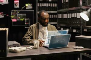 African american policeman working with hologram, planning investigation with holographic evidence and clues in police archive space. Male investigator examining augmented reality photos. photo