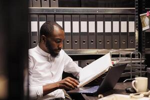 African american prosecutor reading file, preparing for case in police office with archival records. Law enforcement professional analyzing forensic expertise folder at night time photo